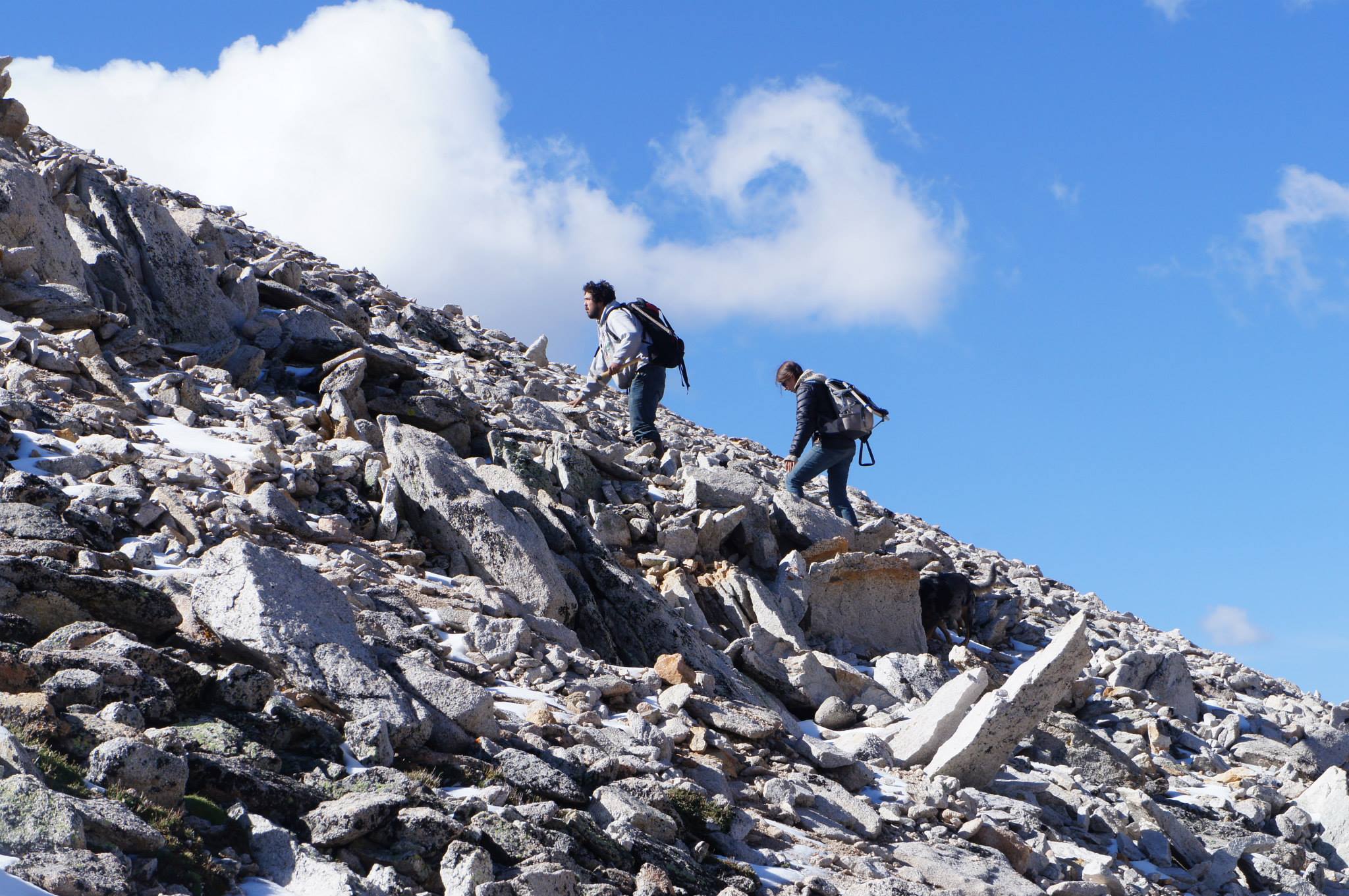 Two people climb a steep mountainside against a blue sky with clouds.